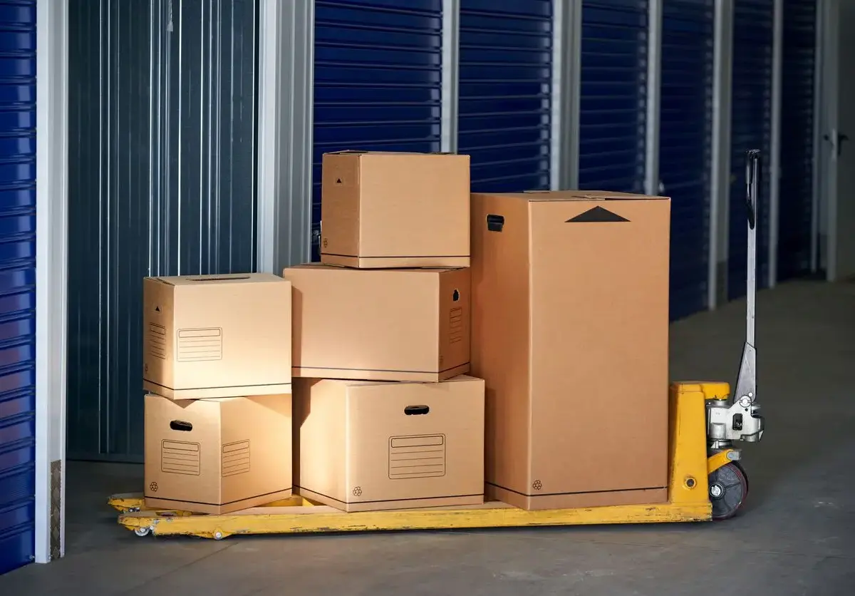 A stack of cardboard boxes on a yellow pallet jack inside a storage facility with blue roll-up doors.