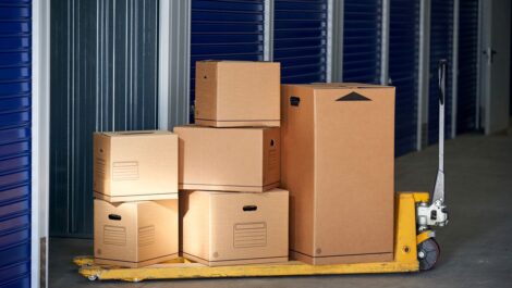 A stack of cardboard boxes on a yellow pallet jack inside a storage facility with blue roll-up doors.