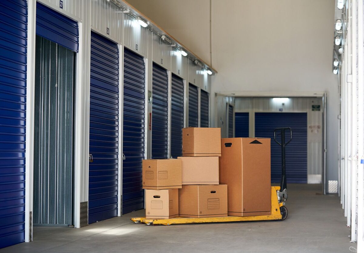 Stacked cardboard boxes on a pallet jack in a clean hallway lined with blue self storage unit doors.