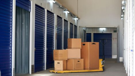 Stacked cardboard boxes on a pallet jack in a clean hallway lined with blue self storage unit doors.